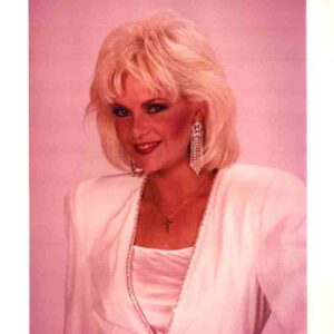 Portrait of a smiling blonde woman in a white blazer with a cross necklace and dangling rhinestone earrings, pink studio backdrop behind her.
