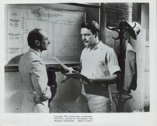 Two men in short-sleeve shirts study a wall chart titled 'Cholera Epidemic' while holding papers.