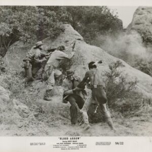 Black-and-white still from Blood Arrow: soldiers and a shirtless fighter taking cover behind rocks as dust rises on a rocky hillside during a firefight.