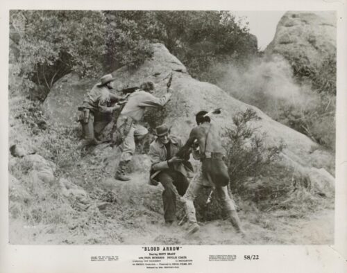 Black-and-white still from Blood Arrow: soldiers and a shirtless fighter taking cover behind rocks as dust rises on a rocky hillside during a firefight.