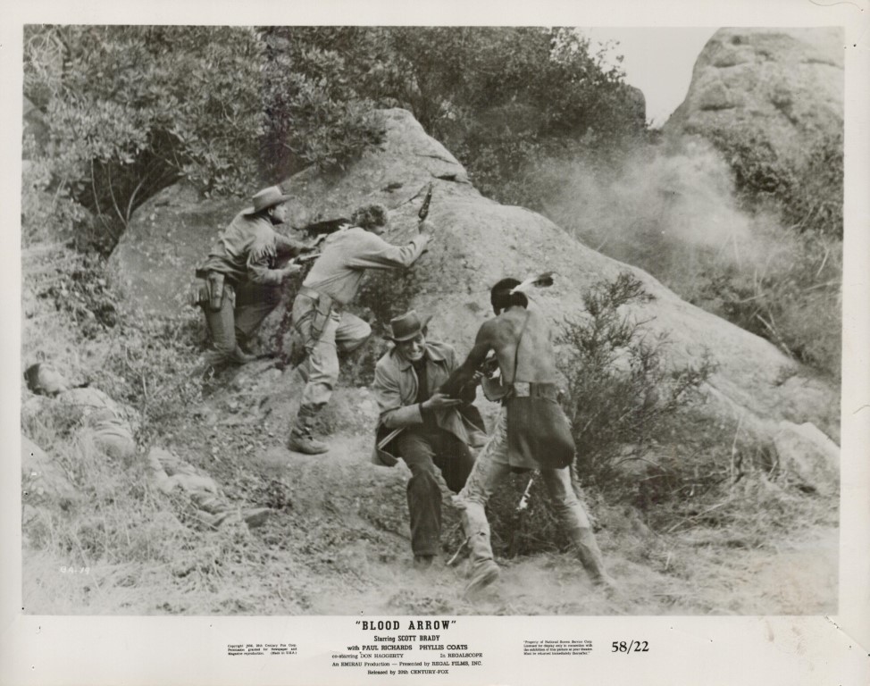 Black-and-white still from Blood Arrow: soldiers and a shirtless fighter taking cover behind rocks as dust rises on a rocky hillside during a firefight.