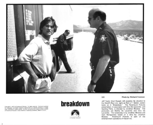 Man in a light polo shirt stands with hands on hips beside a large semi truck on a desert road, facing a uniformed police officer; another man leans on the trailer in the background.