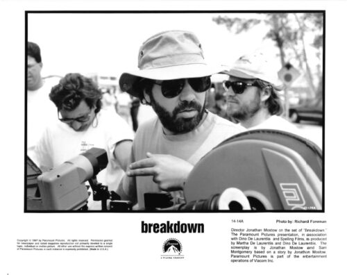 Cameraman wearing hat and sunglasses adjusts equipment on a film set, with others in the background.