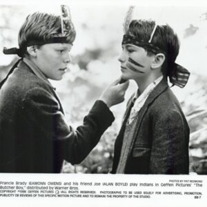 Two boys wearing headbands with feather-like adornments and face paint, posing in a movie still as Indians; one adjusts the other's chin.