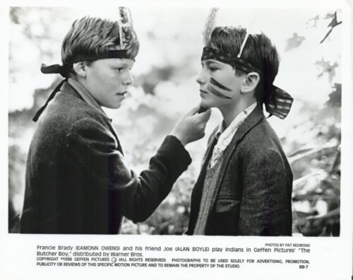 Two boys wearing headbands with feather-like adornments and face paint, posing in a movie still as Indians; one adjusts the other's chin.