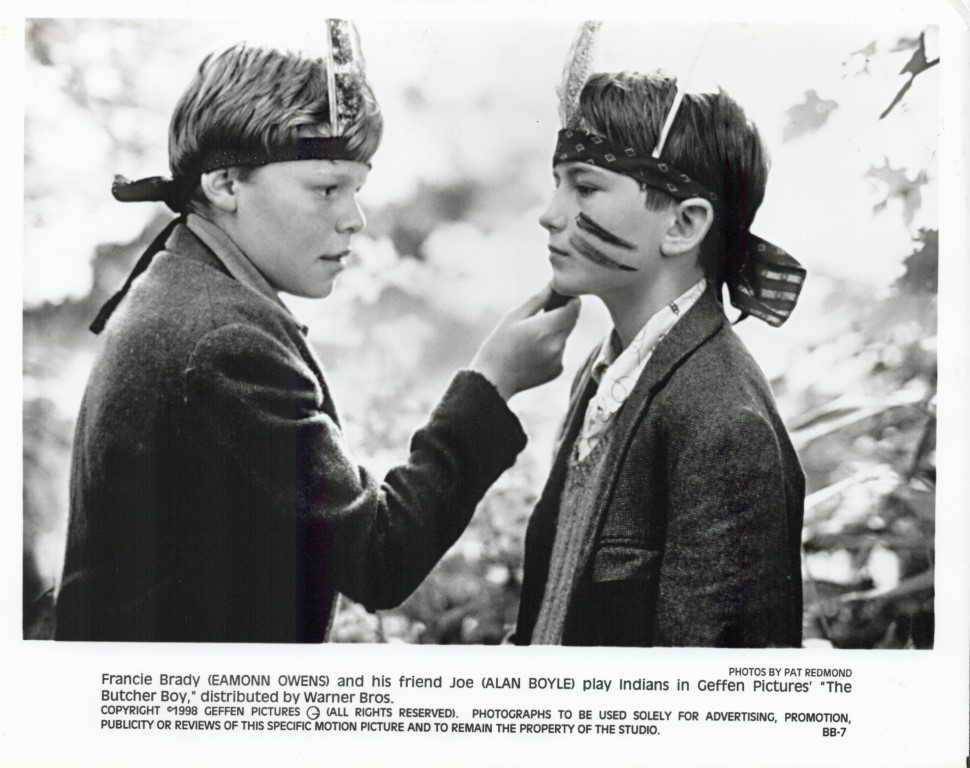 Two boys wearing headbands with feather-like adornments and face paint, posing in a movie still as Indians; one adjusts the other's chin.