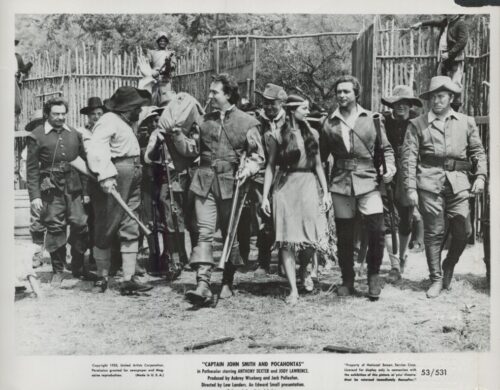 Black-and-white scene from a 1950s film: actors in 17th‑century frontier costumes walk through a rustic outdoor set, led by a man with a rifle and a woman in a fringed dress.