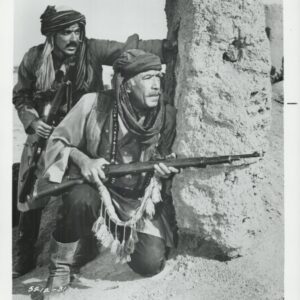 Two armed men in turbans crouch behind a sand wall, looking alert in a desert setting.