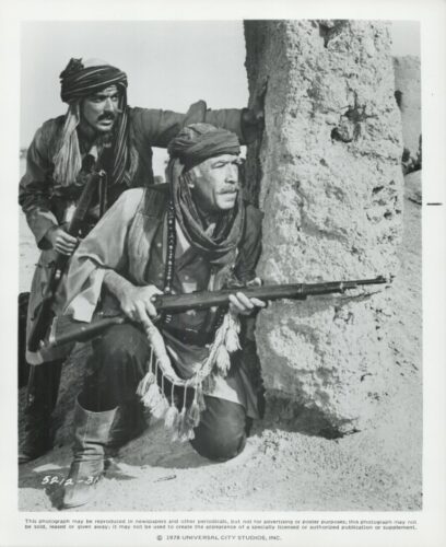 Two armed men in turbans crouch behind a sand wall, looking alert in a desert setting.