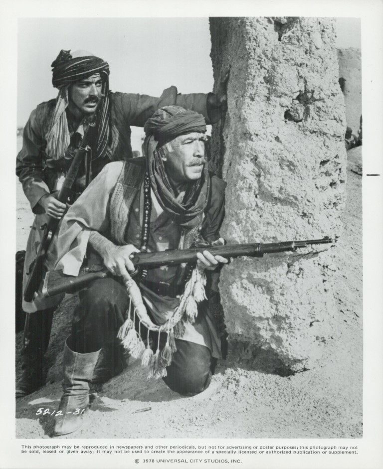 Two armed men in turbans crouch behind a sand wall, looking alert in a desert setting.