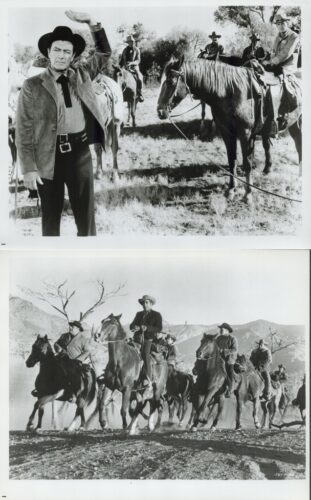 Black-and-white photo of cowboys on horseback; a man in front waves toward the camera while others ride in the background in a prairie setting.