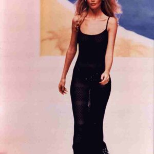 Model walking a runway in a black sheer dress, against a soft blurred backdrop.
