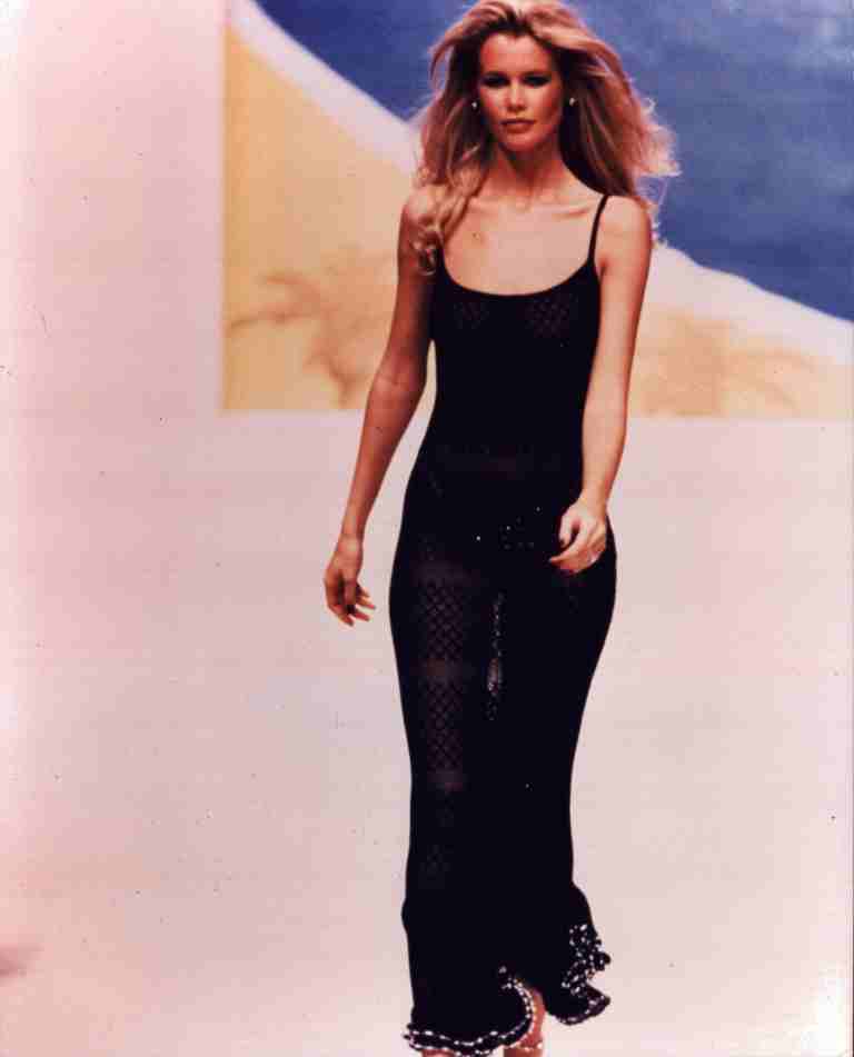 Model walking a runway in a black sheer dress, against a soft blurred backdrop.