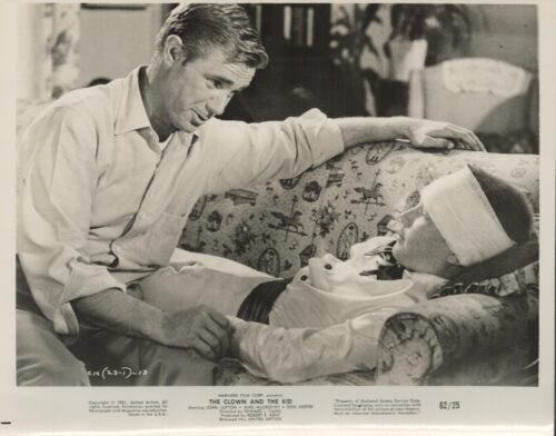 A man sits beside a wounded person with a bandaged head on a sofa, holding their hand in a caring moment.
