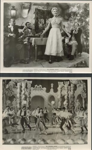 Vintage outdoor scene: a woman in a light dress stands with arms outstretched while four men sit around a table watching her.