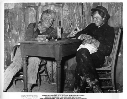 Two men sit at a wooden table in a rough, peeling room, sharing a moment with a bottle and mug on the table between them.