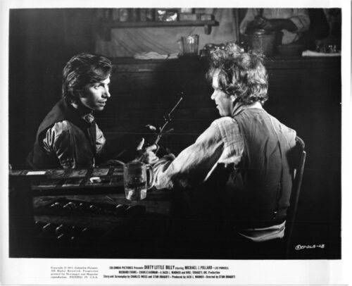 Black-and-white film still: two men sit across from each other at a wooden table, one handing a pistol to the other as they talk.