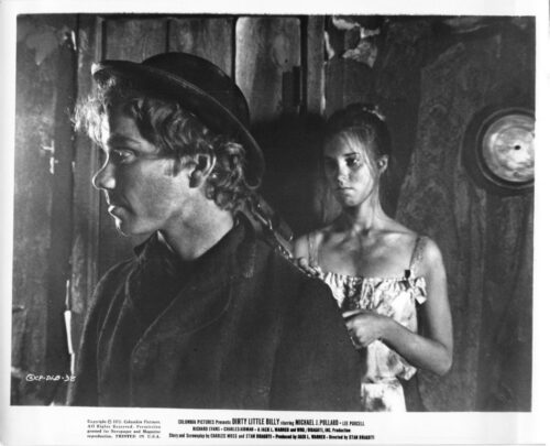 Black-and-white film still: a young man in a wide-brimmed hat and dark jacket is shown in profile to the left, while a woman in an off-shoulder dress stands behind him, her hands near his shoulder in a rustic interior.