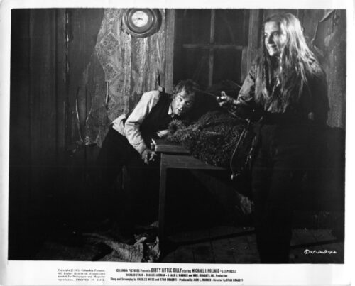 Black-and-white film still from Dirty Little Billy: a man crouches by a table in a dilapidated room as a long-haired woman stands nearby.