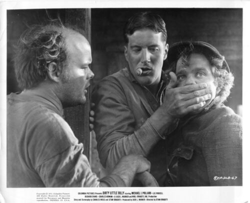 Three rugged men in a doorway; the center man covers the right man's mouth with one hand while smoking a cigar, as the left man looks on.