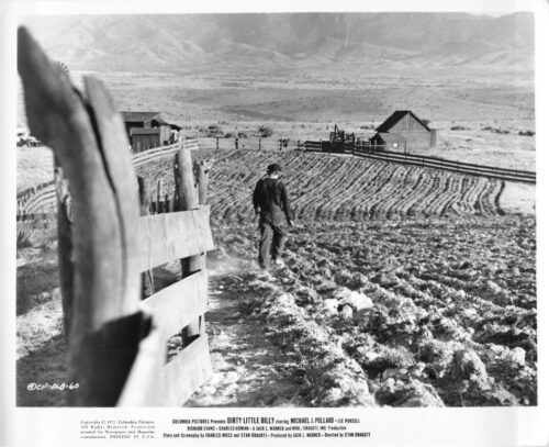 Man walking along a muddy plowed field beside a wooden fence, with barns and mountains in the distance.