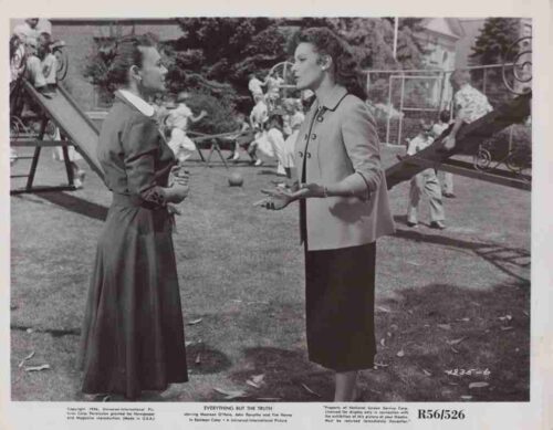 Two women in vintage dresses converse in a park playground, with children on slides and swings in the background.