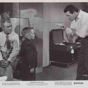 Man in a white shirt points at an open suitcase while a boy and another man watch the scene.