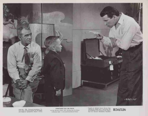 Man in a white shirt points at an open suitcase while a boy and another man watch the scene.