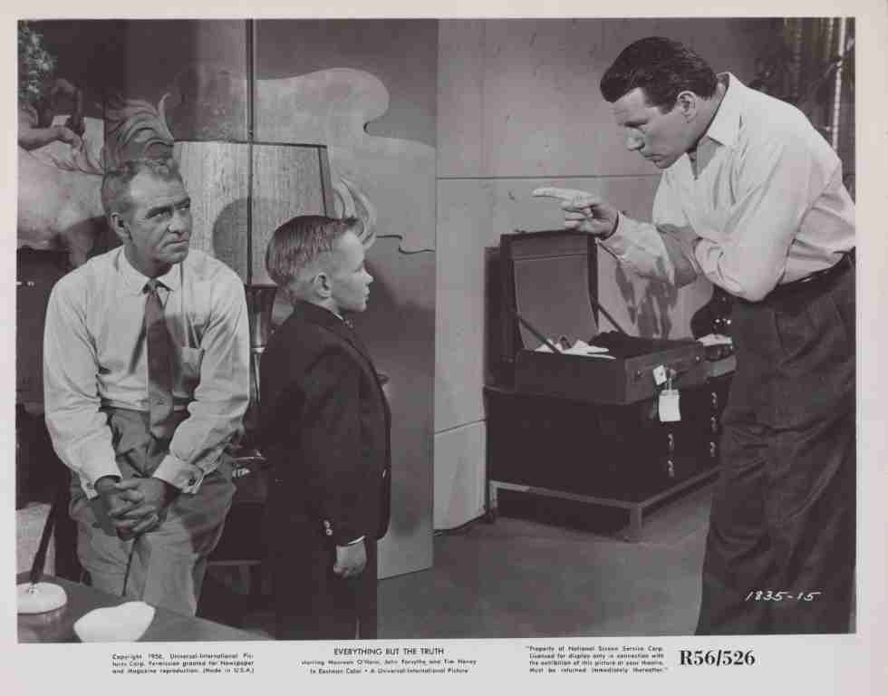 Man in a white shirt points at an open suitcase while a boy and another man watch the scene.