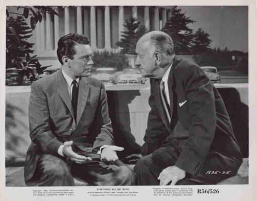 Two men in 1950s suits sit on a low wall, leaning in and talking intently outdoors with cars in the background.