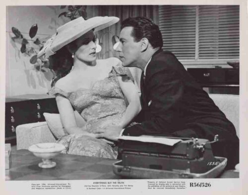 Black-and-white promotional still: a man in a suit leans toward a woman in an off‑the‑shoulder lace dress and wide hat, seated on a couch, with a vintage typewriter on a table in the foreground.