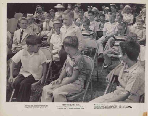 Black-and-white outdoor gathering with children and adults seated on folding chairs, two boys and a girl foreground, spectators in the background.