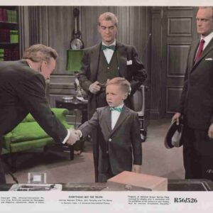 Young boy in a gray suit and green bow tie shakes hands with a man across a desk while three men in suits observe in a vintage office setting.