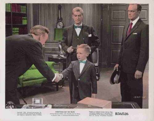 Young boy in a gray suit and green bow tie shakes hands with a man across a desk while three men in suits observe in a vintage office setting.