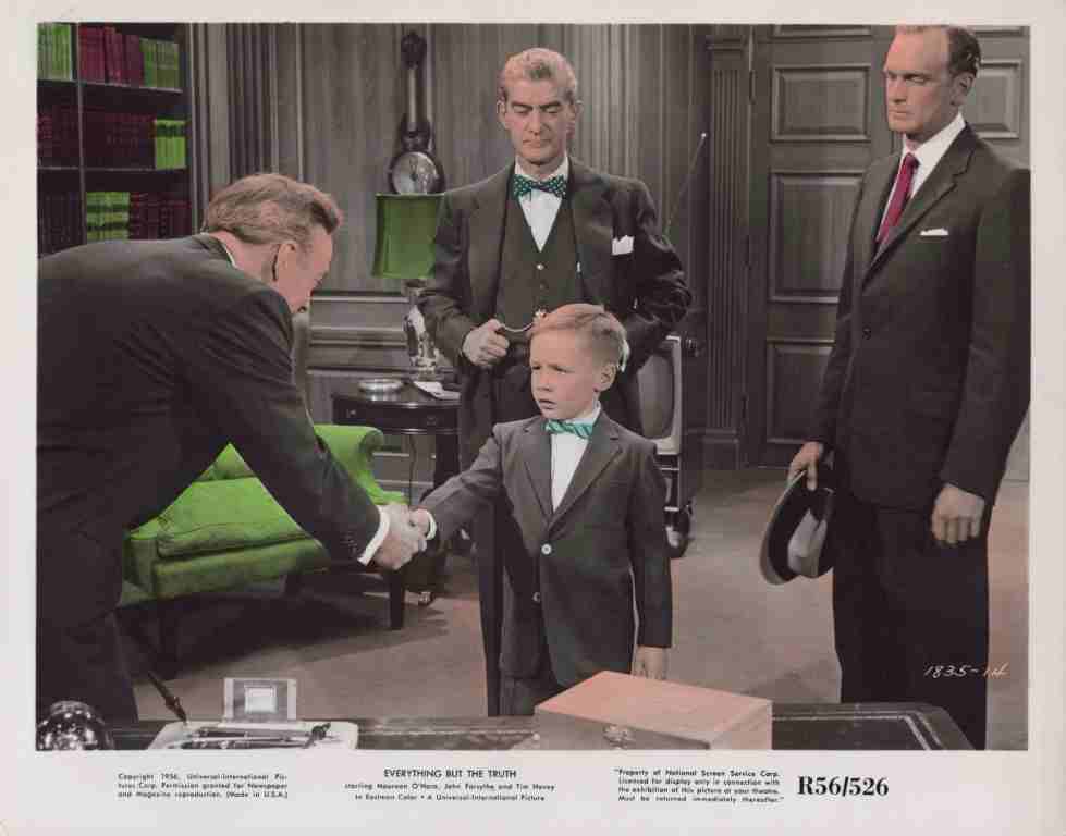 Young boy in a gray suit and green bow tie shakes hands with a man across a desk while three men in suits observe in a vintage office setting.