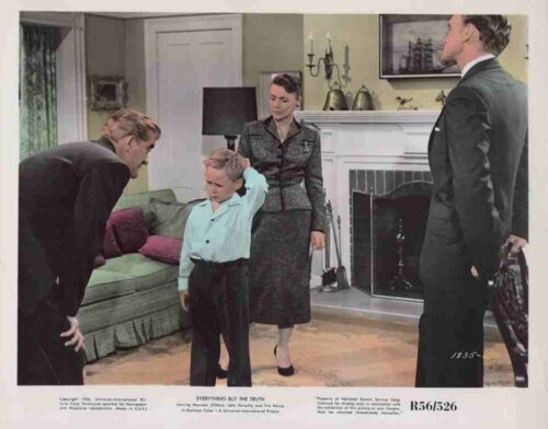 1950s-era film still: a boy in a light blue shirt stands with his hand on his head as three adults in formal attire watch him in a living room with a fireplace and mantle behind them.