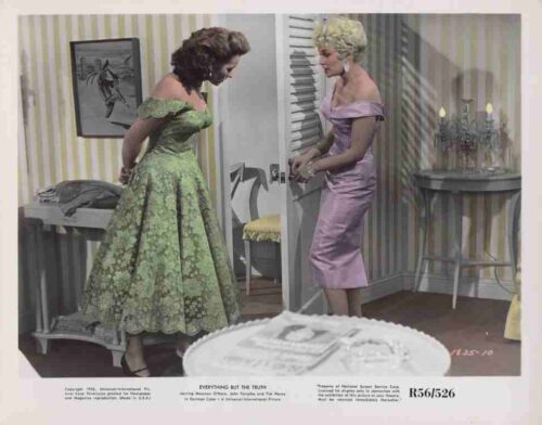 Two women in vintage dresses stand by a doorway, one green lace gown and one lilac satin dress, in a softly lit room with a cake in the foreground.