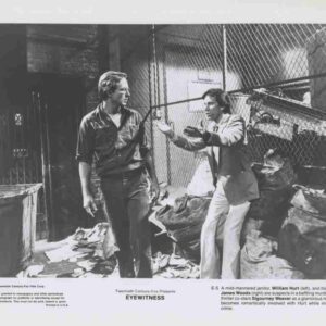 Two men stand in a cluttered warehouse alley, one gesturing toward the other as they converse near a chain-link fence and piles of debris.