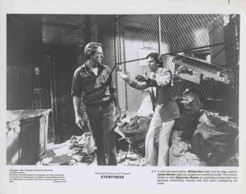 Two men stand in a cluttered warehouse alley, one gesturing toward the other as they converse near a chain-link fence and piles of debris.