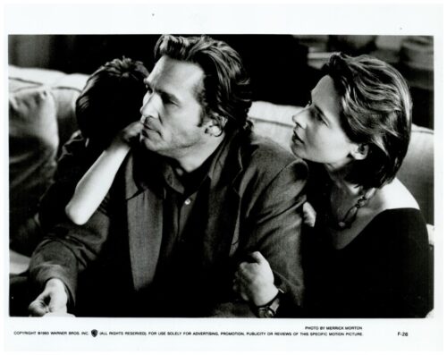 Black-and-white film still: a man in a blazer sits on a couch while a woman leans toward him, her arm around his, both looking off to the left.