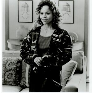 Black-and-white publicity photo of Rosie Perez as Carla in Fearless, standing in a living room, wearing a patterned cardigan over a dark top.