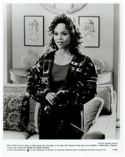 Black-and-white publicity photo of Rosie Perez as Carla in Fearless, standing in a living room, wearing a patterned cardigan over a dark top.