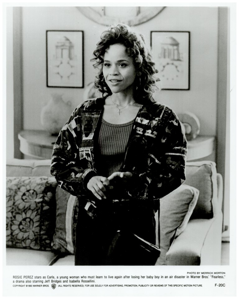 Black-and-white publicity photo of Rosie Perez as Carla in Fearless, standing in a living room, wearing a patterned cardigan over a dark top.