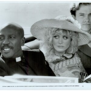 Three adults in a vintage convertible: a smiling priest on the left, a woman in a large hat in the middle, and a suited man in the back.