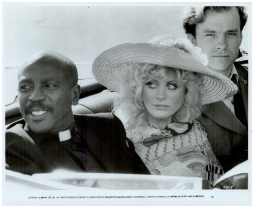 Three adults in a vintage convertible: a smiling priest on the left, a woman in a large hat in the middle, and a suited man in the back.