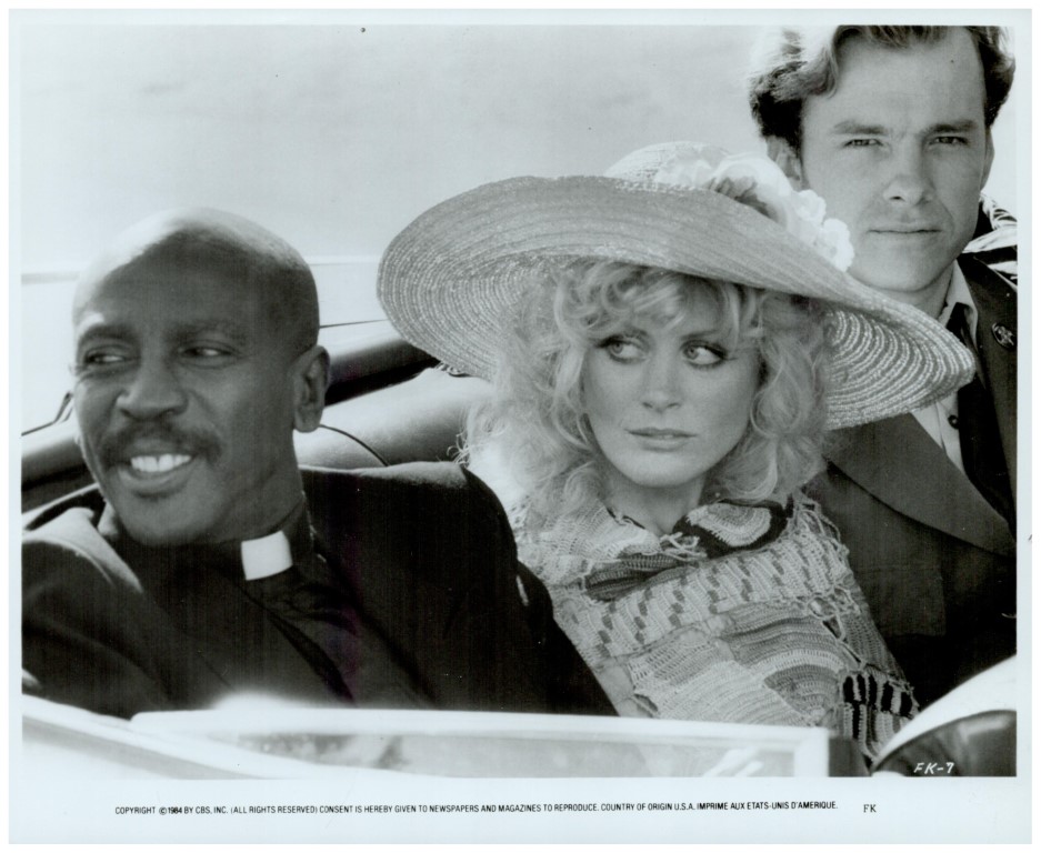 Three adults in a vintage convertible: a smiling priest on the left, a woman in a large hat in the middle, and a suited man in the back.