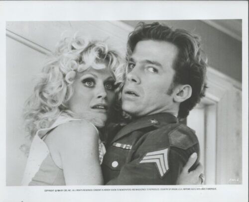 Two people in a tense embrace: a blonde woman clinging to a soldier in a dark uniform, both looking off to the side.