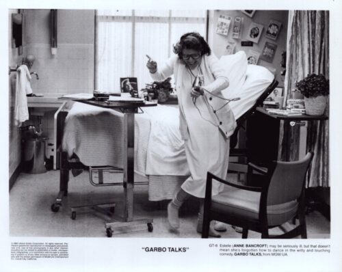 Woman in a white outfit dancing beside a hospital bed, pointing and holding a corded phone in a bright room with a cart and window behind her
