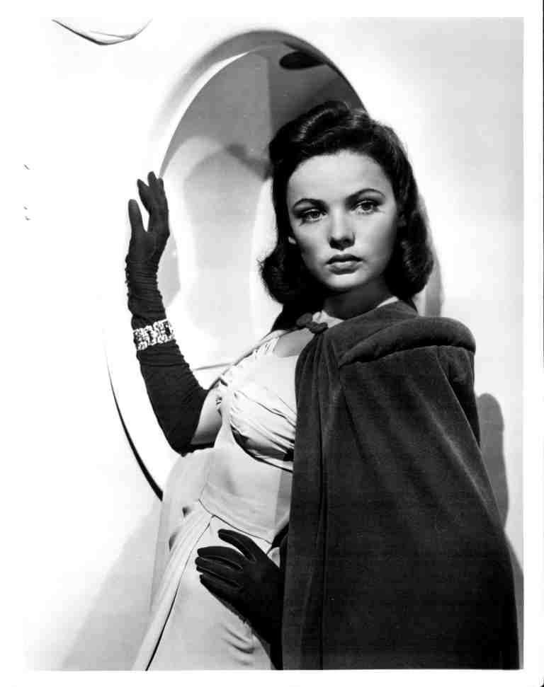 Black-and-white portrait of a glamorous woman in a formal gown and long gloves, posing with a curved backdrop behind her.