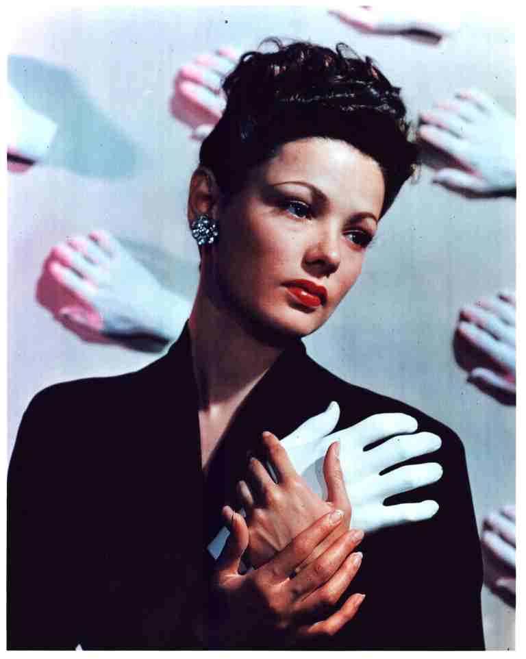 Portrait of a woman in a black coat with short dark hair, looking to the left, surrounded by pale gloved hands in the backdrop.